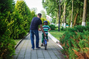 Father help son to ride bicycle in summer park. Back view